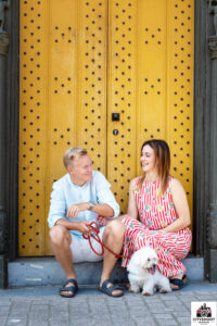 Koppel poseert lachend met hun kleine witte hond voor een opvallende gele houten poort tijdens een Cityshoot in Mechelen door fotograaf Stef Boey.